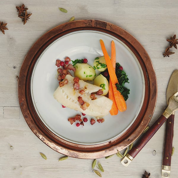 Torsk med bacon, poteter, gulrøtter og grønnkål på tallerken med bestikk på siden